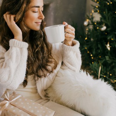 Young woman drinking hot coffee or cocoa sitting on couch in cozy room with Christmas tree. Winter holiday mood.