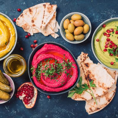 Ingredients for yellow turmeric hummus, green parsley hummus and red beetroot hummus on dark background, lemon, sesame, olive oil, pita, avocado, pomegranate. Middle eastern, jewish, arabic cuisine