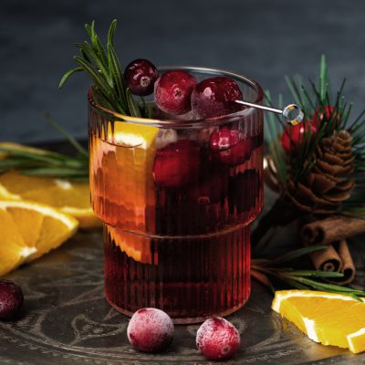 Cranberry boozy Christmas cocktail with ice and rosemary on dark background. Festive spiced drink with orange, berry, cinnamon and anise.